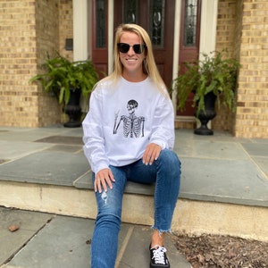 May include: A woman wearing a white long-sleeved sweatshirt with a black skeleton graphic and blue jeans. She is sitting on a set of stone steps outside a house. She is wearing black sunglasses.