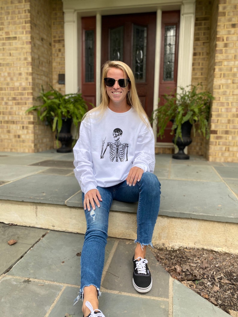 May include: A woman wearing a white long-sleeved sweatshirt with a black skeleton graphic and blue jeans. She is sitting on a set of stone steps outside a house. She is wearing black sunglasses.