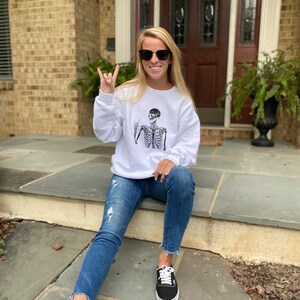 May include: A white sweatshirt with a black skeleton graphic wearing sunglasses and making a rock and roll hand gesture. The person wearing the sweatshirt is sitting on a set of stairs and wearing blue jeans and black sneakers.