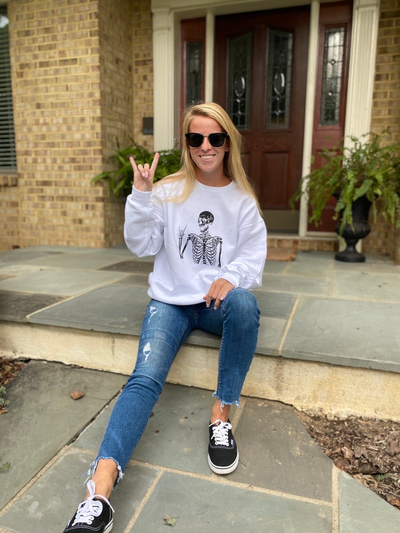May include: A white sweatshirt with a black skeleton graphic wearing sunglasses and making a rock and roll hand gesture. The person wearing the sweatshirt is sitting on a set of stairs and wearing blue jeans and black sneakers.