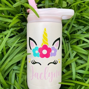 Puede incluir: Una botella de agua de acero inoxidable blanca con una tapa rosa y un dise&ntilde;o de unicornio. El unicornio tiene un cuerno amarillo, orejas rosas y una corona de flores con flores azules, rosas y moradas. El nombre "Jaclynn" est&aacute; escrito en cursiva rosa debajo del unicornio.