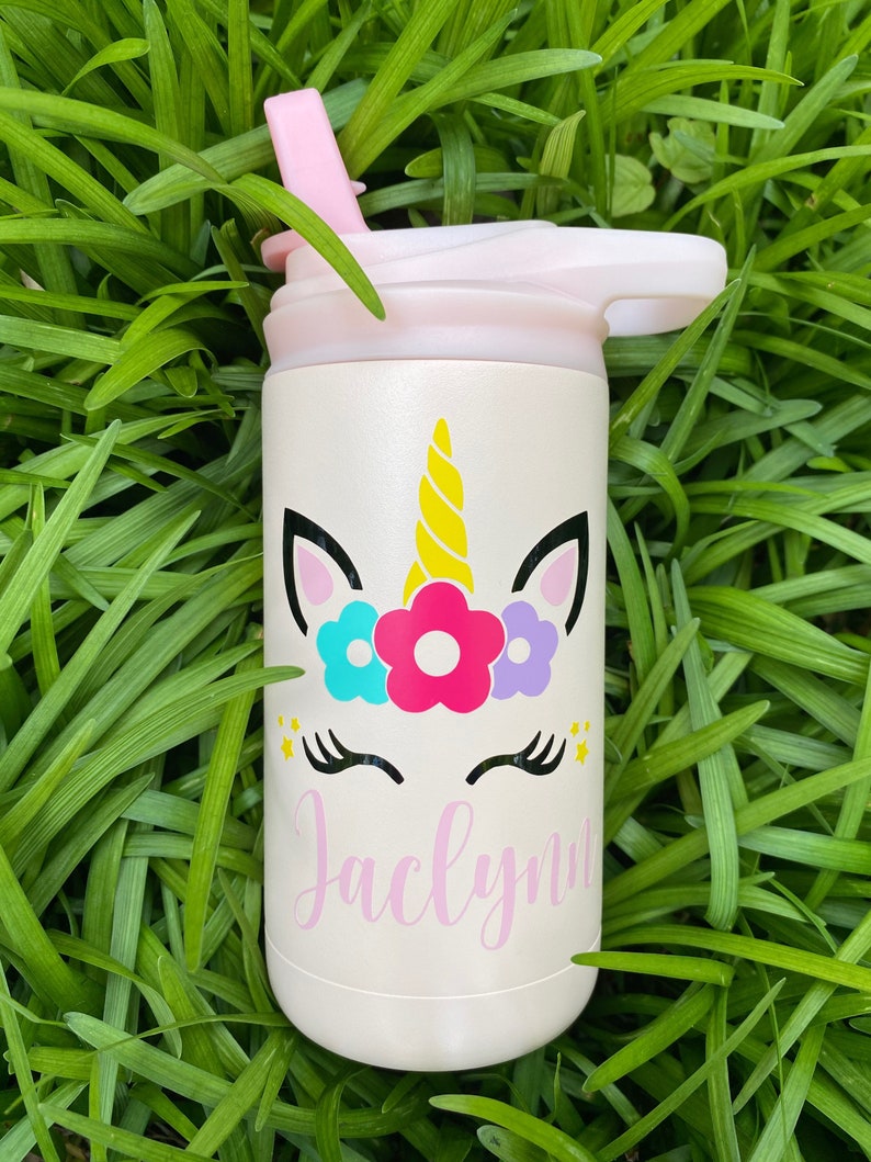Puede incluir: Una botella de agua de acero inoxidable blanca con una tapa rosa y un dise&ntilde;o de unicornio. El unicornio tiene un cuerno amarillo, orejas rosas y una corona de flores con flores azules, rosas y moradas. El nombre "Jaclynn" est&aacute; escrito en cursiva rosa debajo del unicornio.