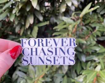 May include: A white sticker with the words "FOREVER CHASING SUNSETS" in navy blue lettering. The sticker is held in front of a background of green leaves.