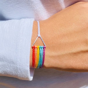May include: A rainbow colored friendship bracelet with a silver triangle charm. The bracelet is made of thin, braided cords.