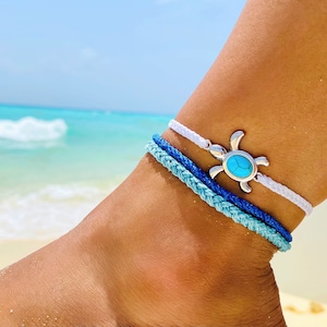 May include: A white and blue braided ankle bracelet with a silver turtle charm featuring a turquoise stone.