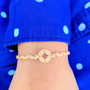 May include: A gold compass charm bracelet with a braided rope band. The compass charm has a star in the center and the letters N, S, E, and W around the edge.