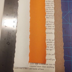 May include: A metal ruler with inch markings rests on a collage of torn paper and book pages. A strip of orange paper is centered between a white torn edge and a book page. The text "ISLAND RESCUE" is visible.