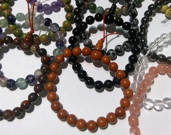 May include: A collection of colorful beaded bracelets made from various natural stones, including brown, black, green, clear, and pink. The bracelets are arranged in a circle on a white surface.