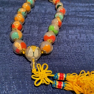May include: A clear glass beaded necklace with colorful painted images inside each bead. The necklace has a yellow tassel with a red and blue tassel attached to it.