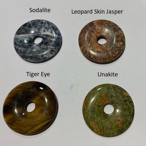 May include: Four round, flat, polished stones with holes in the center. The stones are labeled "Sodalite", "Leopard Skin Jasper", "Tiger Eye", and "Unakite".