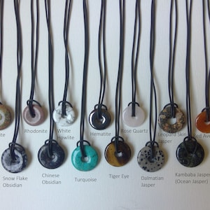 Op de afbeelding: Tien verschillende soorten edelsteen hangers aan zwarte koorden. De hangers zijn rond en hebben een gat in het midden. De edelstenen omvatten aventurijn, sneeuwvlok obsidian, Chinese obsidian, turquoise, tijgeroog, dalmatijnse jaspis, kambaba jaspis, lapis lazuli, unakiet, rhodonite, witte howliet, hematiet, rozenkwarts, luipaardhuid jaspis en rode aventurijn.