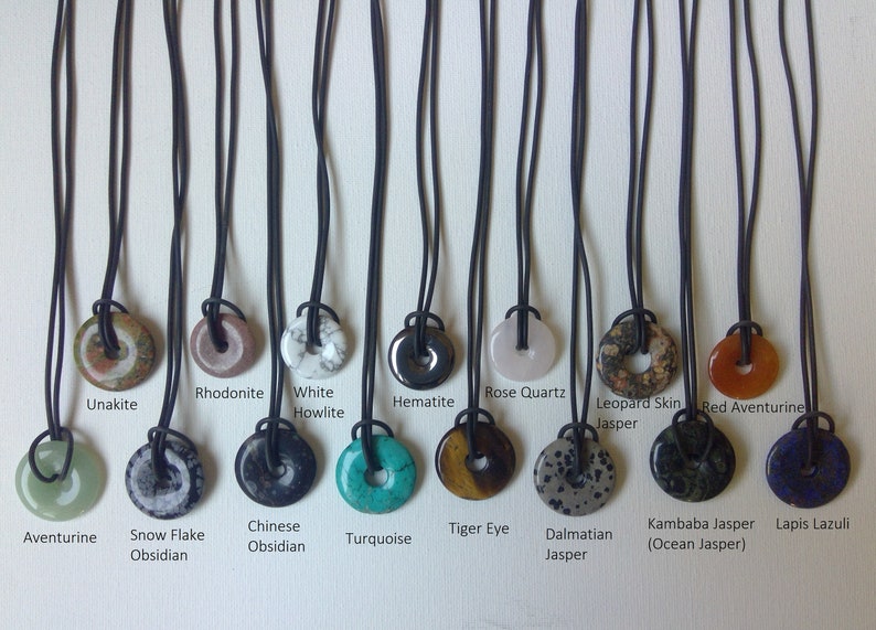 May include: Ten different types of gemstone pendants on black cords. The pendants are round and have a hole in the center. The gemstones include aventurine, snow flake obsidian, Chinese obsidian, turquoise, tiger eye, dalmatian jasper, kambaba jasper, lapis lazuli, unakite, rhodonite, white howlite, hematite, rose quartz, leopard skin jasper, and red aventurine.