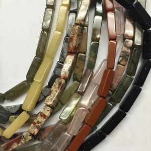 May include: A collection of colorful, rectangular gemstone beads. The beads are made of various types of stone, including green, brown, red, black, and white. The beads are arranged in a loose pile, and some of them are overlapping.