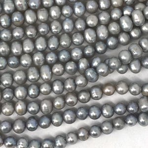 Pode incluir: Uma coleção de numerosas pequenas pérolas de água doce redondas e cinzentas dispostas em várias filas paralelas. Cada pérola apresenta uma superfície lisa e brilhante com variações subtis de tom e forma, criando um aspeto cinzento metálico uniforme.