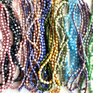 May include: Multiple strands of small, oval-shaped glass beads in a variety of colors including pink, black, blue, orange, yellow, green, and purple. Each bead features a distinct, fine vertical striped pattern running along its length.