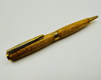 May include: A wooden pen with gold-colored accents. The pen features a light brown, textured body with a gold clip and tip. Gold bands encircle the pen near the center. The pen is designed for writing.