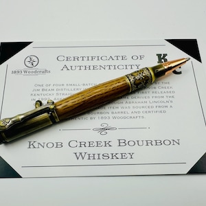 May include: A certificate of authenticity for Knob Creek Bourbon Whiskey. The certificate is printed on white paper with black text and a gold seal. A wooden pen with a gold-plated tip and a brass cap is lying on top of the certificate. The pen is made from a bourbon barrel.