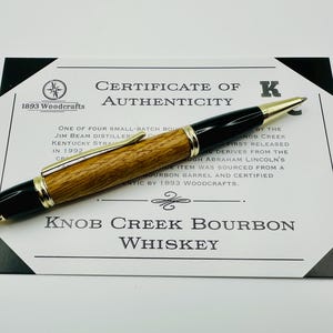 May include: A wooden pen with a black and gold trim and a certificate of authenticity. The certificate states that the pen is made from a bourbon barrel sourced from Knob Creek Distillery. The certificate is signed by 1893 Woodcrafts.