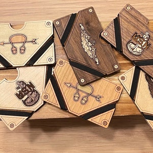 May include: Wooden wallets with various designs, including an anchor, a ship, and an arrow with a loop. The wallets are made of light and dark wood, with black elastic bands and metal screws. The wallets are a unique accessory.