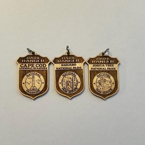 May include: Three wooden keychains with the text "Hark Ranger" and the names of three national parks: Cape Cod National Seashore, Saguaro National Park, and Joshua Tree National Park. Each keychain features a small image of a landmark from the park.
