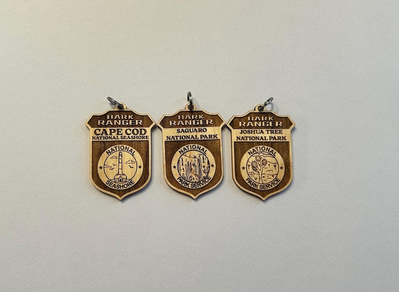 May include: Three wooden keychains with the text "Hark Ranger" and the names of three national parks: Cape Cod National Seashore, Saguaro National Park, and Joshua Tree National Park. Each keychain features a small image of a landmark from the park.