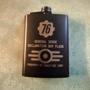 May include: Black rectangular flask with a screw-on cap. The flask features the number "76" inside a circular design, along with the text "GENERAL ISSUE RECLAMATION DAY FLASK" and "PROPERTY OF VAULT-TEC CORP" in silver lettering.