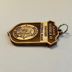 May include: Wooden keychain with a brown and tan color scheme. It features a park ranger badge with the text "Bark Ranger" and "Saguaro National Park".