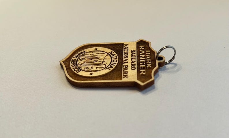 May include: Wooden keychain with a brown and tan color scheme. It features a park ranger badge with the text "Bark Ranger" and "Saguaro National Park".
