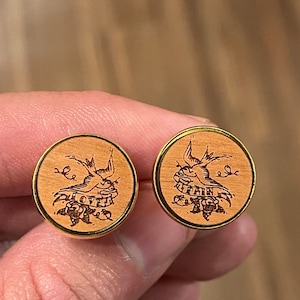 May include: A pair of wooden cufflinks with a gold trim. Each cufflink features a laser-engraved design of a bird with a banner that reads "Love is True".