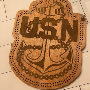 May include: A wooden game board with a US Navy anchor and the letters USN. The board has a circular shape with holes around the edge.