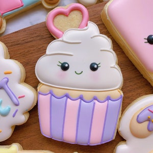 May include: A decorated cupcake cookie with a smiling face, white frosting, and a pink heart on top. The cupcake has a purple and pink striped base. Other decorated cookies are in the background.