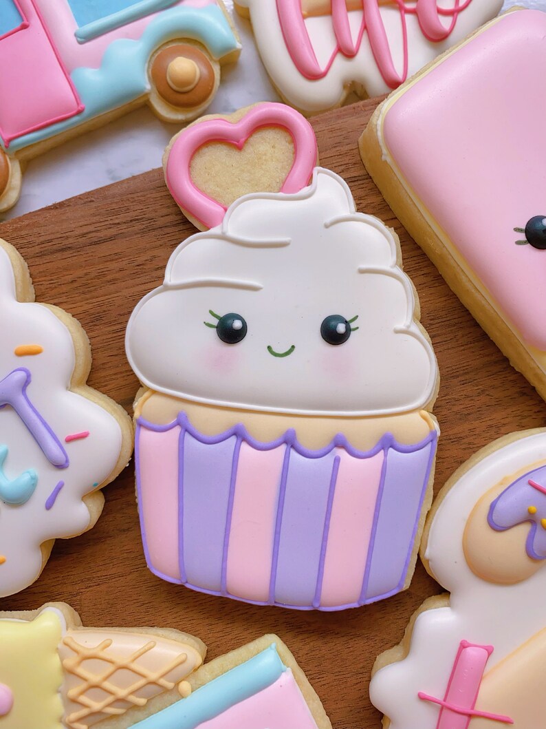May include: A decorated cupcake cookie with a smiling face, white frosting, and a pink heart on top. The cupcake has a purple and pink striped base. Other decorated cookies are in the background.