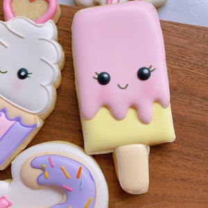 May include: A collection of decorated sugar cookies. The cookies include a pink popsicle with a smiling face, a cupcake, a donut with sprinkles, and a cookie with the word "two" in pink icing.