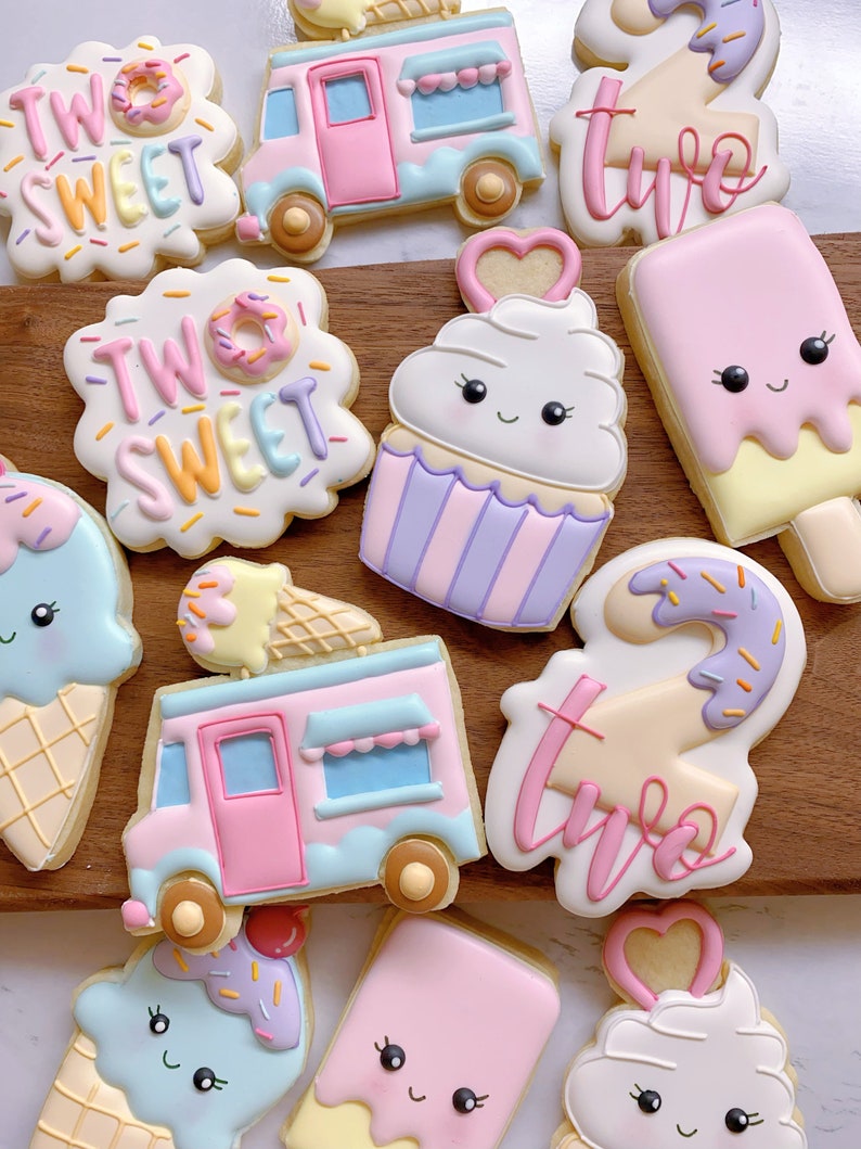 May include: Assortment of decorated sugar cookies in the shape of ice cream cones, ice cream bars, a cupcake, a food truck, and cookies with the text "Two Sweet". The cookies are decorated with pastel colors and sprinkles.