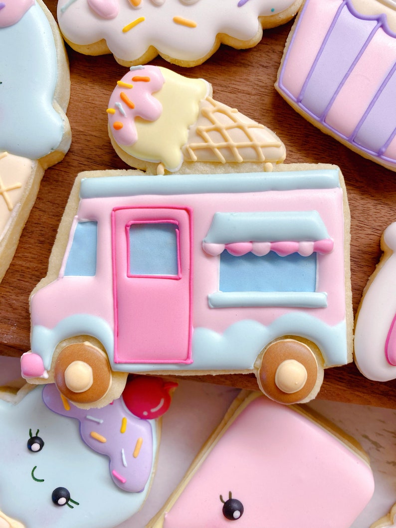 May include: A collection of decorated sugar cookies in pastel colors. The cookies include an ice cream truck, an ice cream cone, and other shapes. The ice cream truck cookie is pink and blue with brown wheels. The ice cream cone has yellow ice cream with sprinkles.