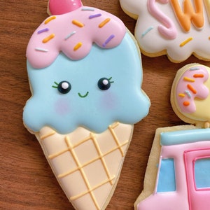 May include: A decorated sugar cookie shaped like an ice cream cone. The ice cream is light blue with pink sprinkles and a pink cherry on top. The cone is beige with a waffle pattern. Other cookies are visible.