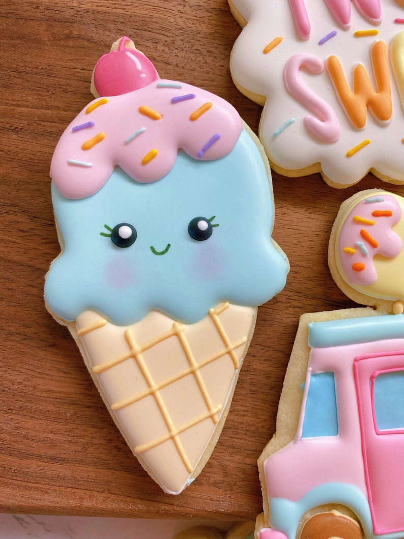 May include: A decorated sugar cookie shaped like an ice cream cone. The ice cream is light blue with pink sprinkles and a pink cherry on top. The cone is beige with a waffle pattern. Other cookies are visible.