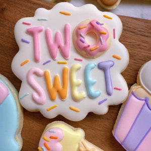 May include: A decorated sugar cookie with the words "TWO SWEET" in pastel colors, with a donut in place of the "O". The cookie is white with colorful sprinkles. Other cookies in the image include an ice cream cone and a vehicle.
