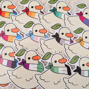 Puede incluir: Una colección de pegatinas con patos de dibujos animados con picos naranjas y pañuelos con diseños de banderas del orgullo. Cada pato tiene una hoja verde en la cabeza. Las pegatinas están sobre un fondo blanco.