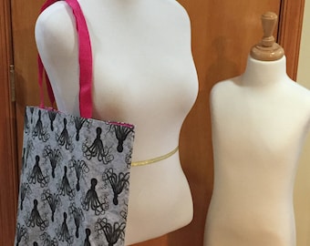 Könnte beinhalten: Eine graue Einkaufstasche mit einem rosa Griff und einem schwarzen und weißen Kraken-Druck. Die Tasche befindet sich auf einem weißen Mannequin.