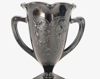 Peut inclure: Un vase en verre noir avec deux anses, un motif en forme de cœur et une base carrée. Le vase est décoré d'un motif en relief de deux personnages se tenant la main.