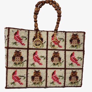 May include: A rectangular tote bag with a beaded wooden handle. The bag's body is a cream-colored needlepoint with a grid pattern featuring red cardinals and brown owls. Gold-colored rings connect the handle to the bag.