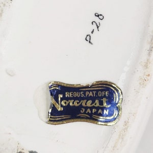 May include: A blue and gold label with the text "Reg. U.S. Pat. Off. Norcrest Japan" on a white ceramic surface.