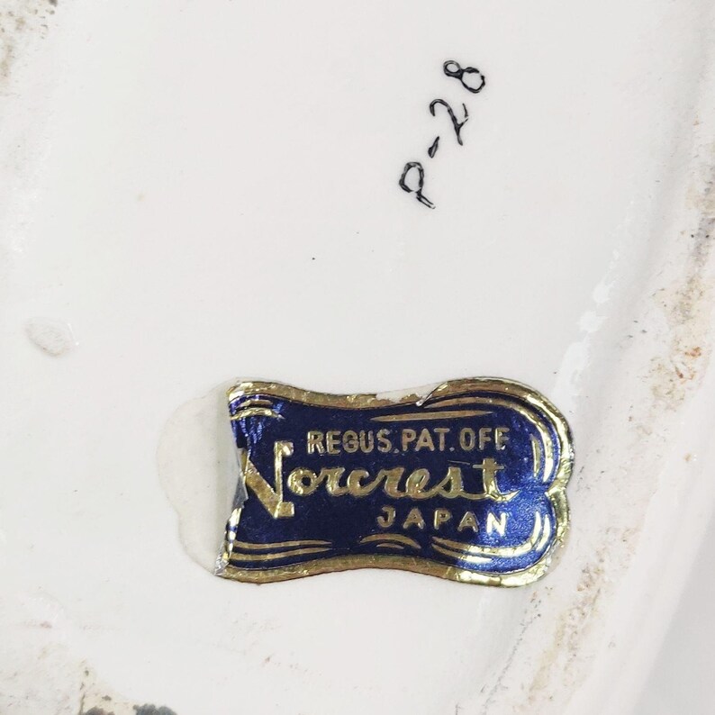 May include: A blue and gold label with the text "Reg. U.S. Pat. Off. Norcrest Japan" on a white ceramic surface.