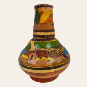 Puede incluir: Un jarrón de cerámica pintado a mano con un diseño marrón, amarillo, verde y azul. El jarrón presenta una escena de una casa con árboles y una cerca.