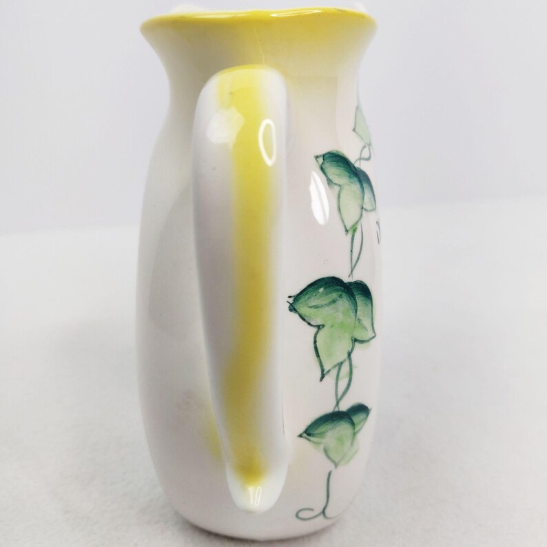 May include: White ceramic creamer with a yellow handle and green ivy design.
