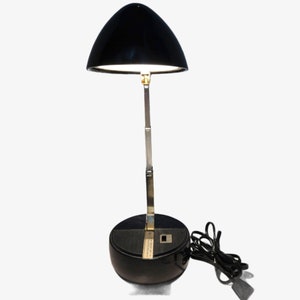 May include: A black desk lamp with a curved shade and a silver-colored adjustable arm. The lamp has a round base and a power cord. The shade is angled downward, and the light is on.