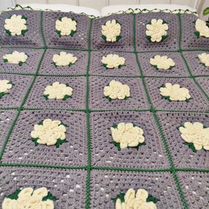 Puede incluir: Una manta de crochet gris con un patrón repetitivo de flores amarillas. La manta está hecha con un punto de granny square y tiene un borde verde.