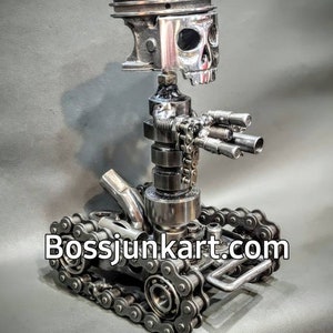 May include: A metal sculpture of a skull with a gun and tank treads. The sculpture is made from recycled metal parts, including a piston, chains, and bolts. The skull is silver and the tank treads are black. The sculpture is signed "Bossjunkart.com".