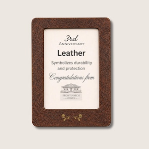 Puede incluir: Un marco de fotos rectangular con un acabado de cuero marrón texturizado. El inserto blanco presenta texto negro que dice 3rd Anniversary, Leather, Symbolizes durability and protection, Congratulations from y un gráfico de una casa Front Porch Homes.